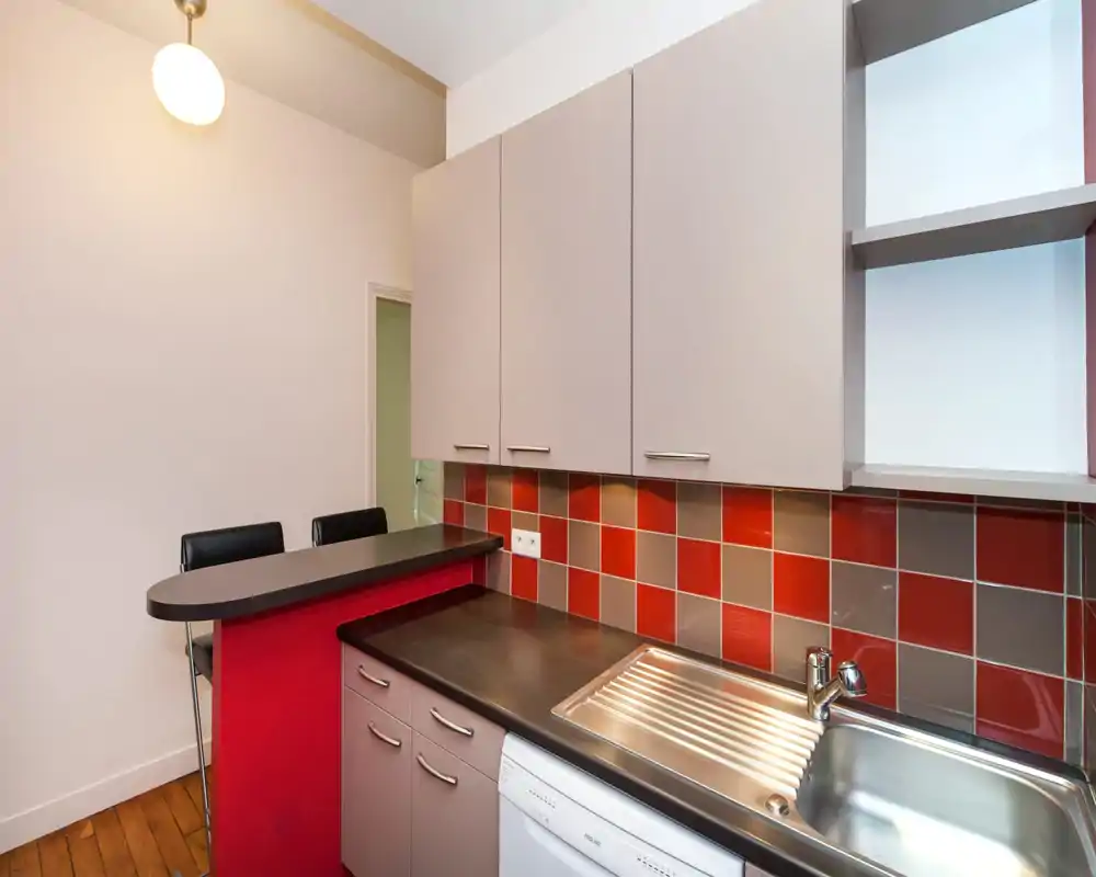 Cucina compatta e moderna con paraschizzi a piastrelle rosse e grigie, piano scuro, lavello in acciaio inox e mobile incassato — perfetta per un appartamento piccolo.