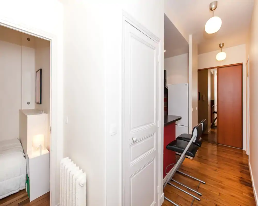 Camera da letto piccola visibile attraverso una porta aperta con letto parzialmente visibile, biancheria bianca e una lampada da comodino; in primo piano corridoio e angolo cucina.