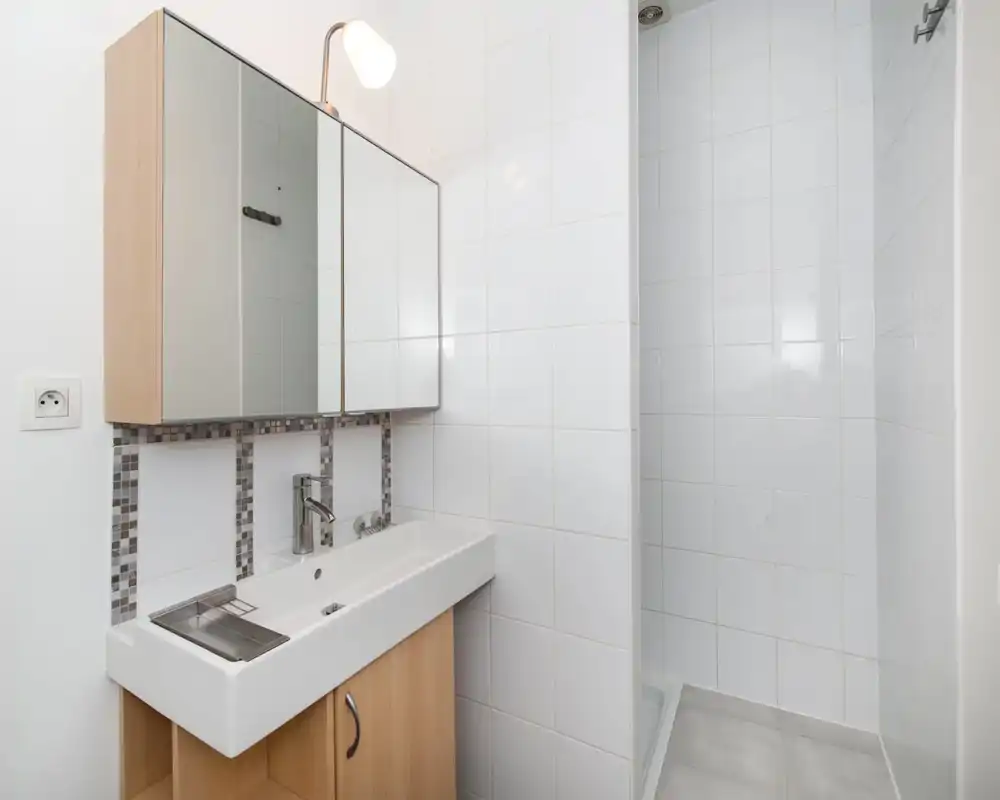 Bagno compatto e moderno con pensile da parete con specchio, lavabo rettangolare con rubinetto cromato e zona doccia piastrellata. Piastrelle bianche e mobili in legno chiaro creano un ambiente luminoso e minimale.