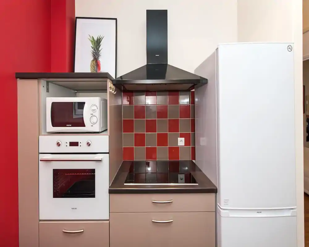 Angolo cucina moderno e compatto con forno incassato, microonde, cappa aspirante e frigorifero, rivestimento a piastrelle rosse e marroni e piano di lavoro pulito.