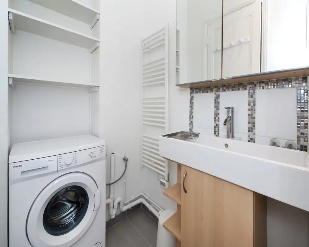 Bagno compatto con lavanderia integrata: lavabo bianco con specchiera, piastrelle decorative e lavatrice a carica frontale sotto ripiani—spazio luminoso e funzionale.