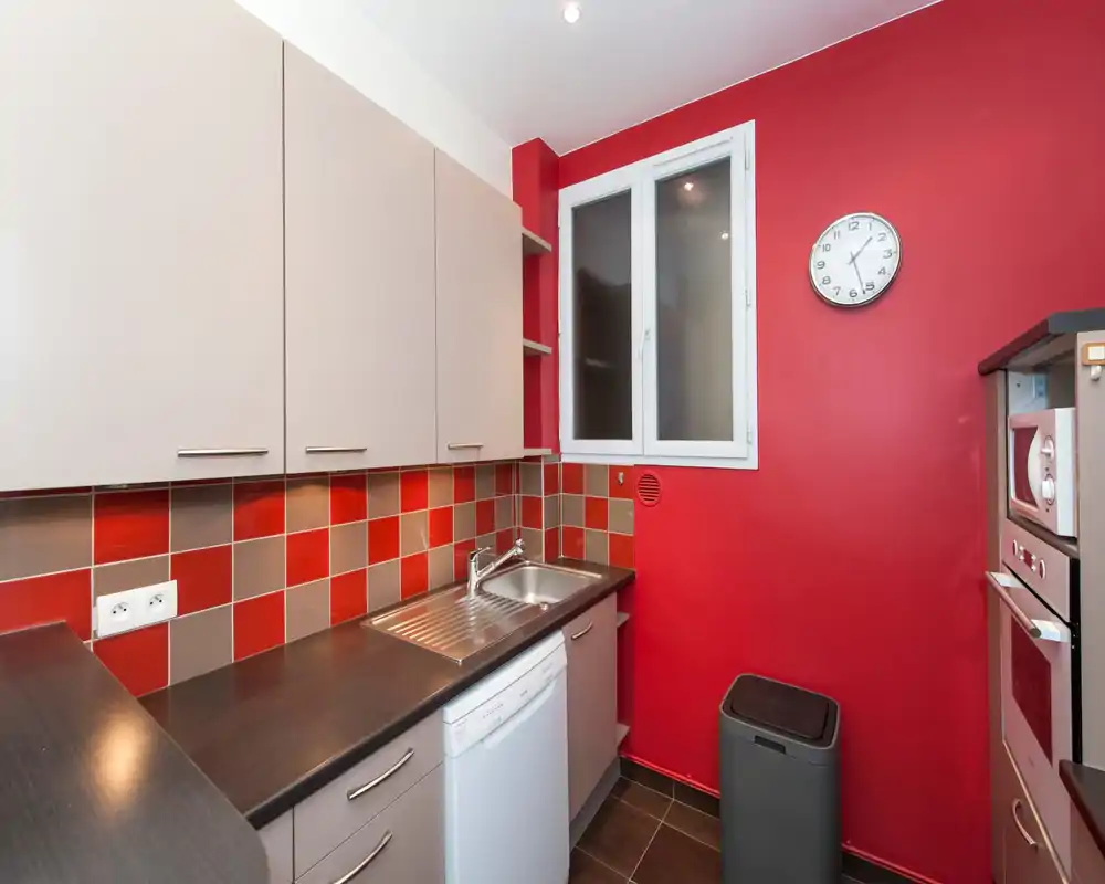 Cucina moderna e compatta con rivestimento a piastrelle rosse e beige, lavello integrato e lavastoviglie, e pensili coordinati. Layout luminoso e funzionale, ideale per piccoli appartamenti.