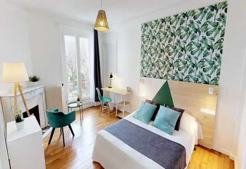 Camera da letto luminosa e di stile con letto matrimoniale, testiera decorata a foglie, pavimento in legno e scrivania vicino alla finestra — perfetta per rilassarsi o lavorare da remoto.