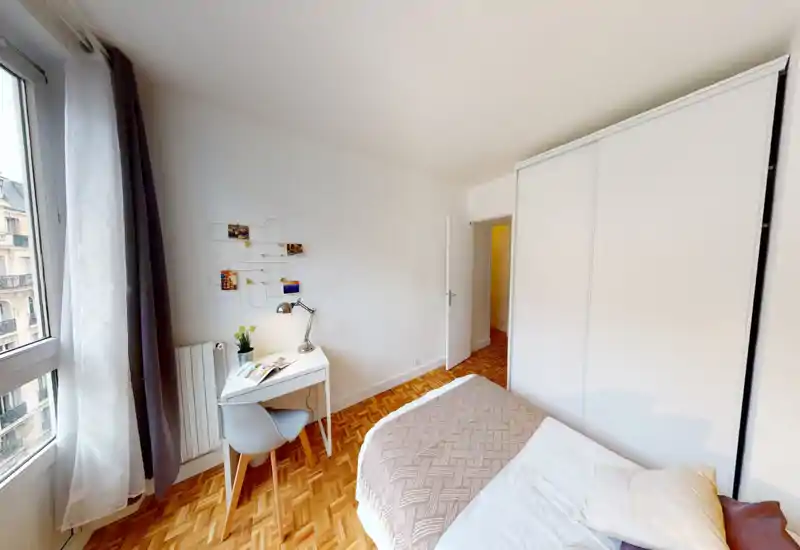 Camera da letto luminosa e accogliente con letto comodo, coperta decorativa e una postazione di lavoro accanto a una grande finestra che illumina la stanza.