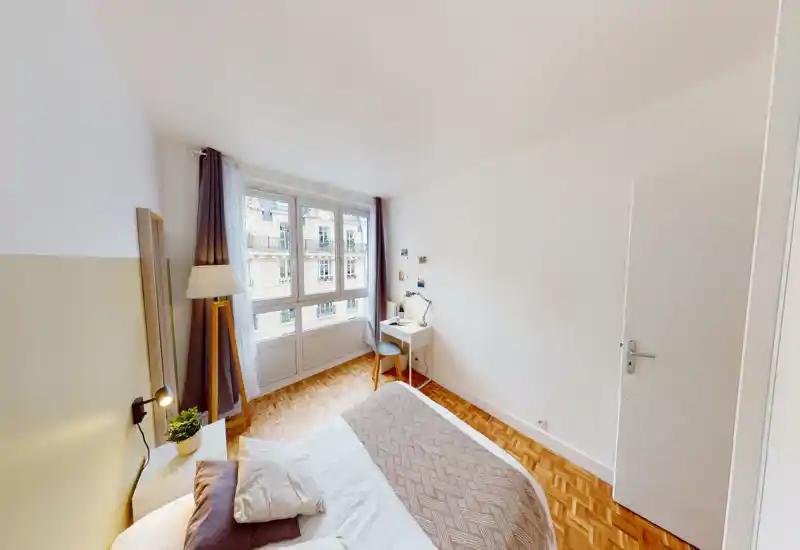 Camera da letto luminosa e compatta con letto matrimoniale vicino a una grande finestra. Pavimento in parquet caldo, lampada da comodino e una piccola scrivania perfetta per studiare o lavorare.
