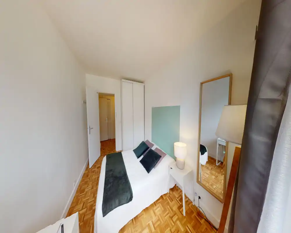 Bright, neatly styled bedroom with a double bed, decorative cushions and a bedside lamp. Hardwood parquet and a full-length mirror add warmth and depth.