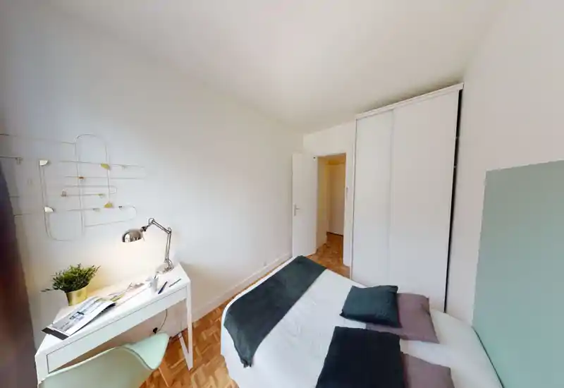 Bright, modern bedroom with a neatly made double bed, cozy pillows and a decorative throw. A small desk with lamp and plant sits beside the bed and a built-in wardrobe provides storage.