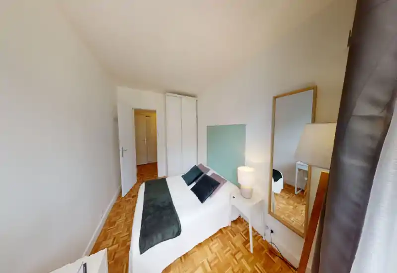 Bright, neatly styled bedroom with a double bed, decorative cushions and a bedside lamp. Hardwood parquet and a full-length mirror add warmth and depth.