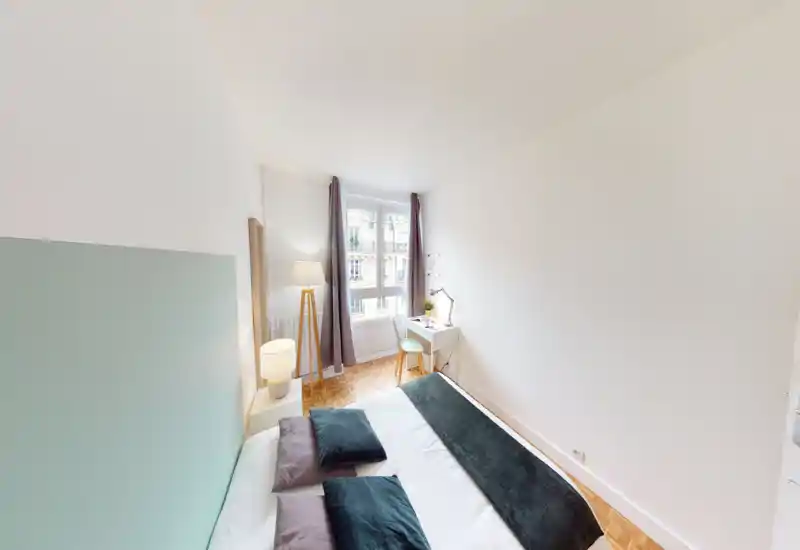Light, compact bedroom with a neatly made double bed, soft cushions, a floor lamp and a small desk by the window. Bright natural light and minimalist décor create a calm, modern atmosphere ideal for short or long stays.