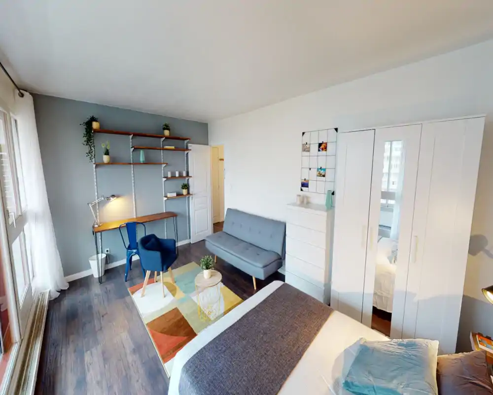 Dormitorio moderno y luminoso con gran ventanal, cama bien hecha en primer plano, sofá compacto, escritorio y estanterías—ideal para una estancia cómoda.