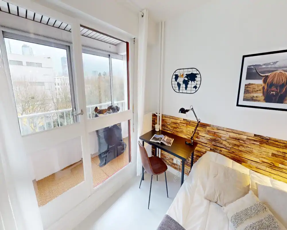 Chambre lumineuse et compacte avec un lit double, un petit bureau et un accès au balcon par de grandes baies vitrées. Tête de lit en bois et textiles doux offrent une ambiance chaleureuse et moderne.