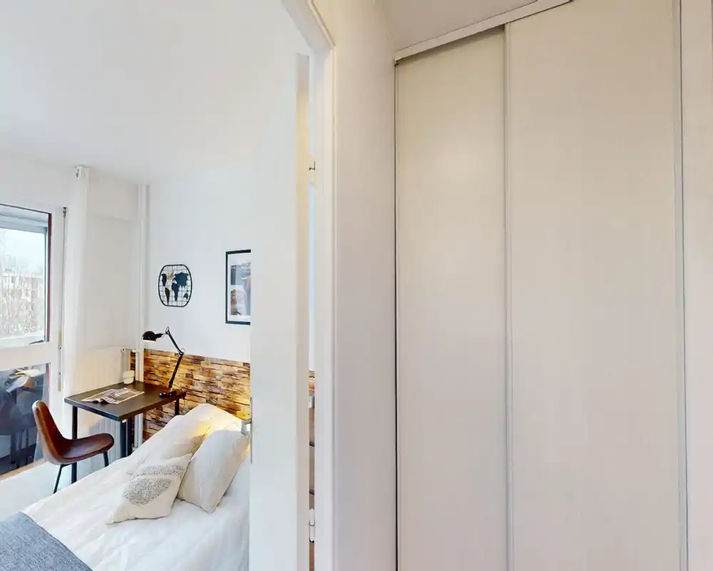 Chambre lumineuse et bien rangée avec un lit double, une tête de lit texturée et un petit bureau près de la fenêtre — idéale pour une personne seule ou un couple ; grande fenêtre apportant une belle lumière naturelle.