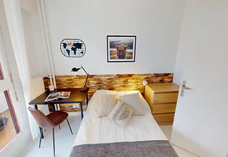 Chambre lumineuse et chaleureuse avec lit double, coussins décoratifs et parement mural en bois. Un petit bureau avec lampe se trouve à côté du lit et une commode offre du rangement — parfaite pour un séjour confortable.