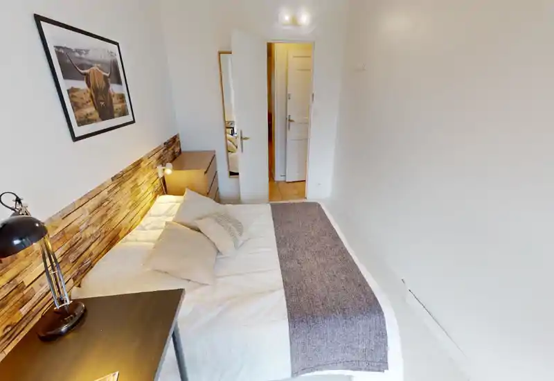 Chambre compacte et bien aménagée avec lit double, tête de lit aspect bois, lampe de chevet et petit bureau — ambiance lumineuse et chaleureuse, parfaite pour de courts séjours.