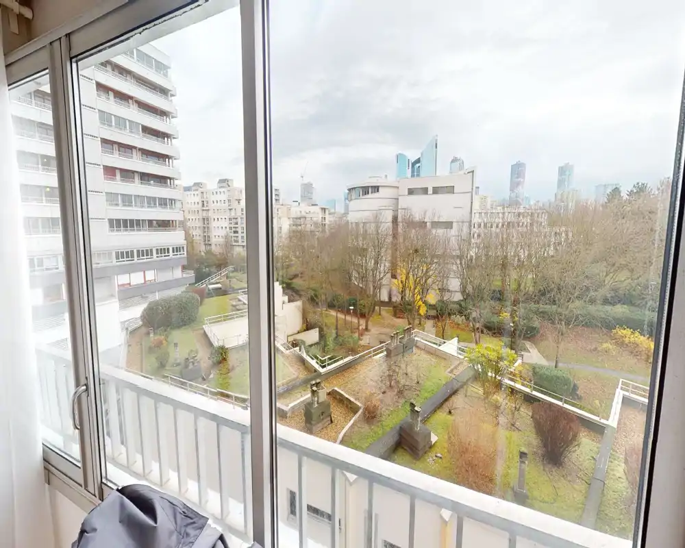 Vue depuis le balcon sur une cour paysagée et les immeubles environnants, avec de grandes baies vitrées laissant entrer beaucoup de lumière naturelle.