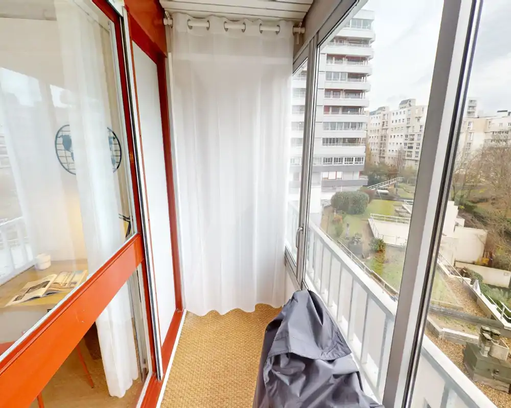 Balcon fermé et lumineux avec grandes baies vitrées donnant sur une cour résidentielle ; sol en fibre et rideaux clairs créent un espace extérieur abrité.