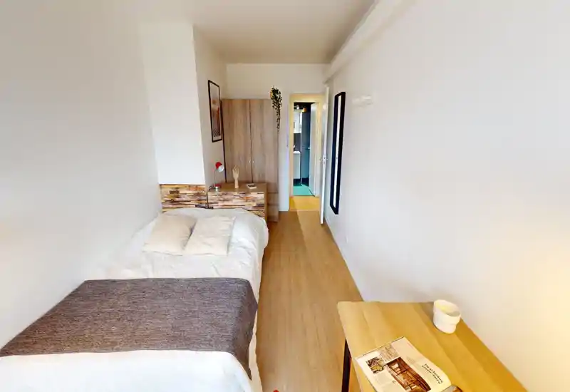 Bright, well-staged bedroom with a double bed, soft linens and a wood-style headboard. A wardrobe and nightstand sit at the foot of the bed and a small desk is visible in the foreground — a cozy, minimal space ideal for short stays.