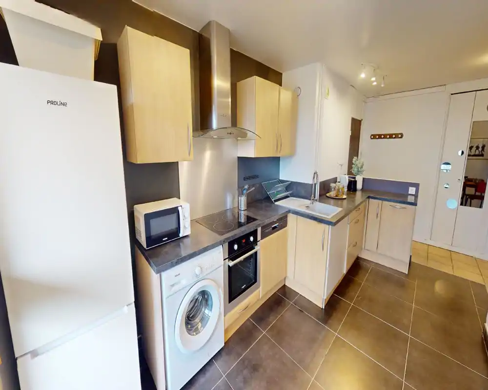 Cuisine moderne équipée avec placards en bois clair, plaque vitrocéramique, four encastré et lave-linge ; plans de travail lumineux et évier intégré adaptés à un usage quotidien.