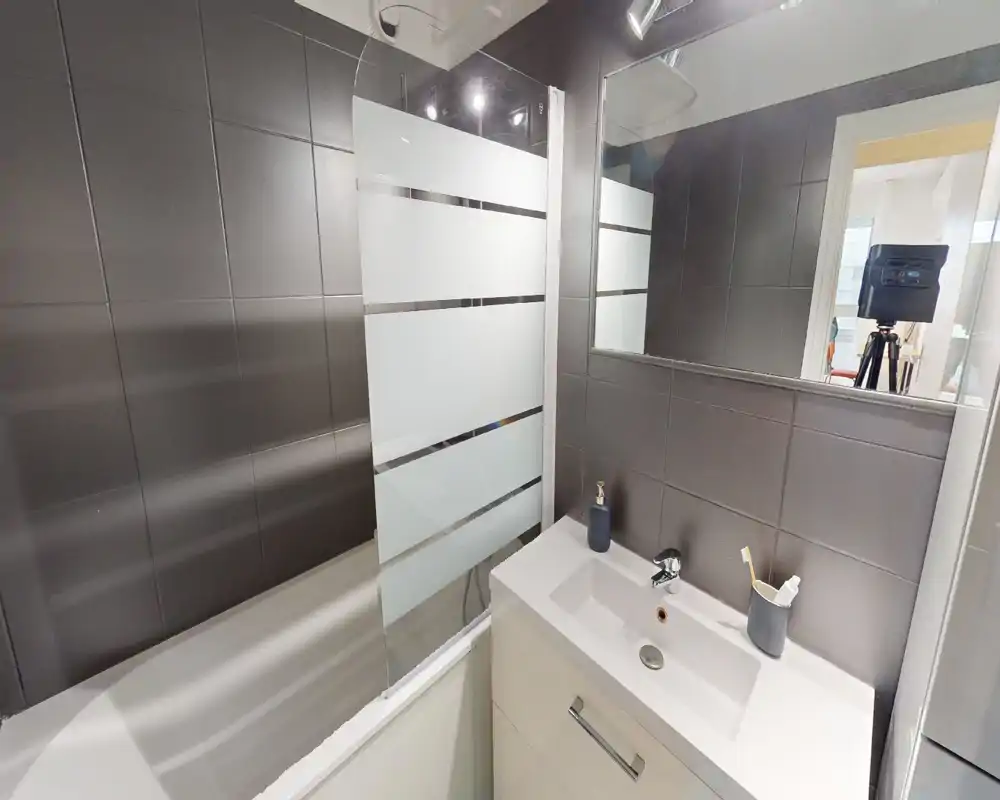 Salle de bains moderne avec carrelage foncé, baignoire avec pare-douche en verre dépoli et lavabo blanc avec meuble; agencement lumineux, propre et fonctionnel, idéal pour une annonce.