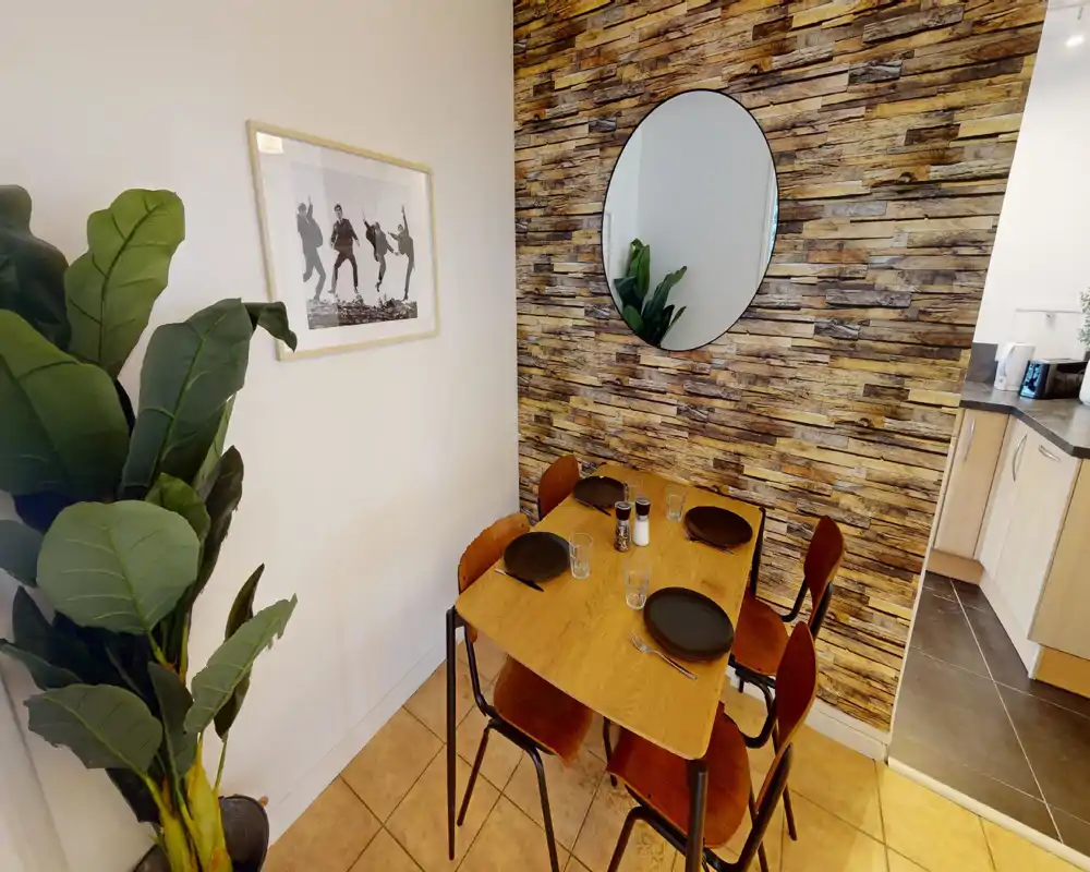 Coin repas chaleureux avec une table en bois pour quatre, un mur effet bois texturé et un miroir rond qui apporte de la profondeur. Lumineux et bien décoré, il communique avec la cuisine pour des repas conviviaux.