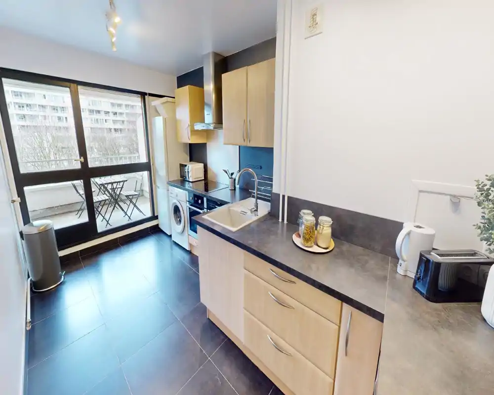 Cuisine moderne et lumineuse avec évier, petits appareils, four et lave-linge ; grandes baies vitrées donnant sur un balcon avec table et chaises.