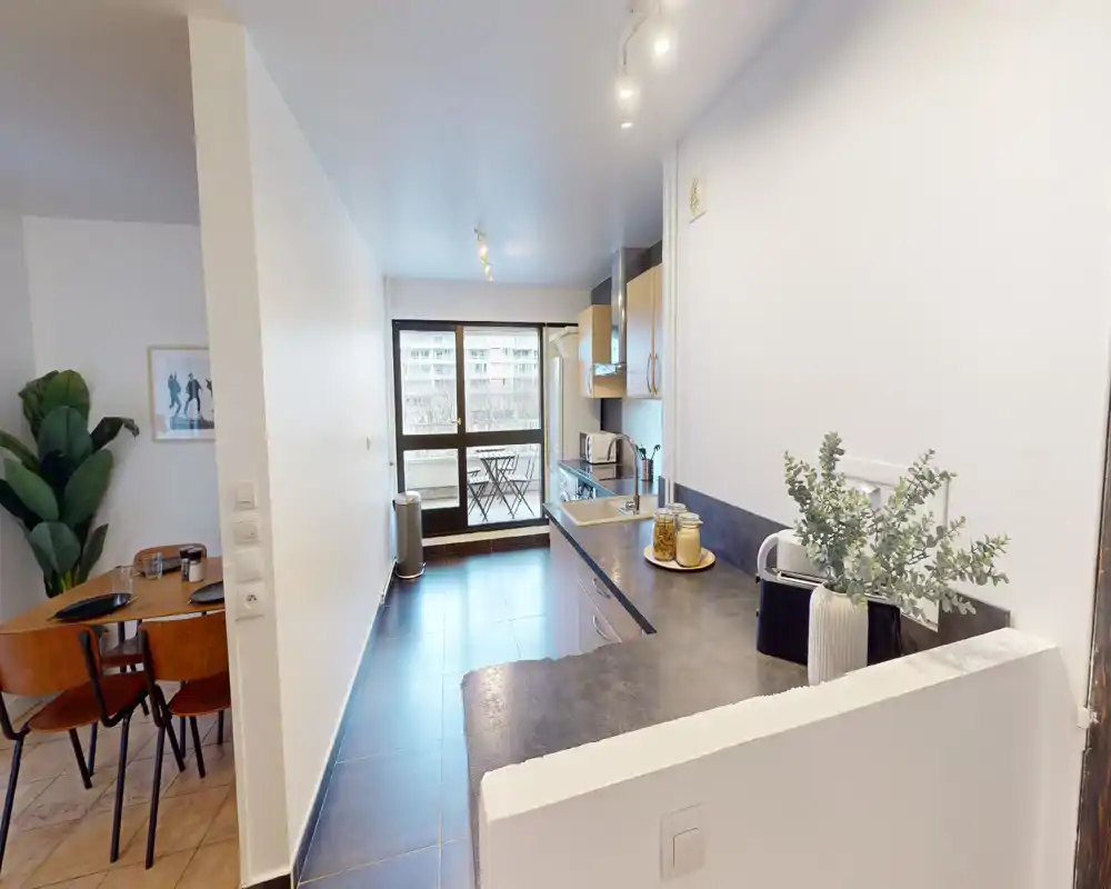 Cuisine moderne ouverte avec un long plan de travail, évier intégré et appareils, donnant sur un balcon avec coin repas extérieur. Lignes épurées, tons neutres et décoration soignée pour un espace de cuisson lumineux et fonctionnel.