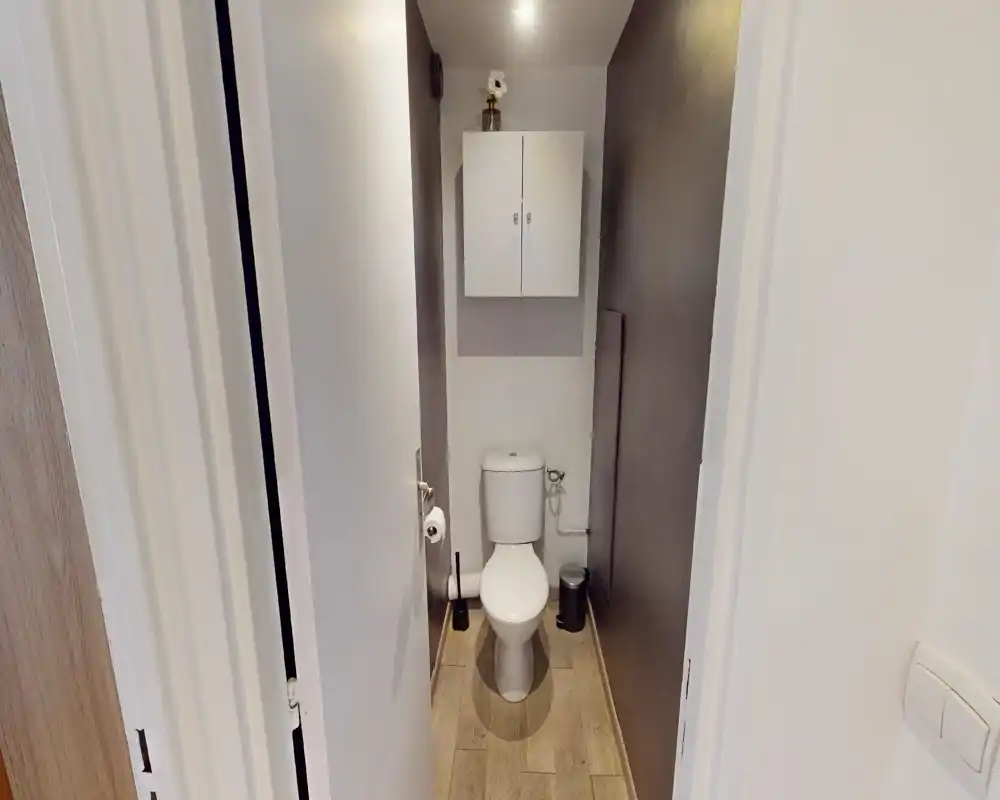 Toilettes compactes et bien éclairées avec placard mural, toilette en céramique blanche, brosse et petite poubelle ; décoration neutre gris et blanc avec sol effet bois.