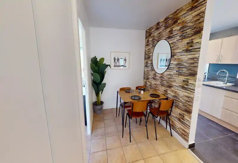 Compact dining area with a wooden table for four, warm textured accent wall and a round mirror; adjacent to the kitchen for easy access.