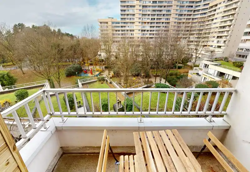 Balcony with a small wooden table and chairs overlooking a landscaped courtyard and apartment buildings — bright view ideal for morning coffee.