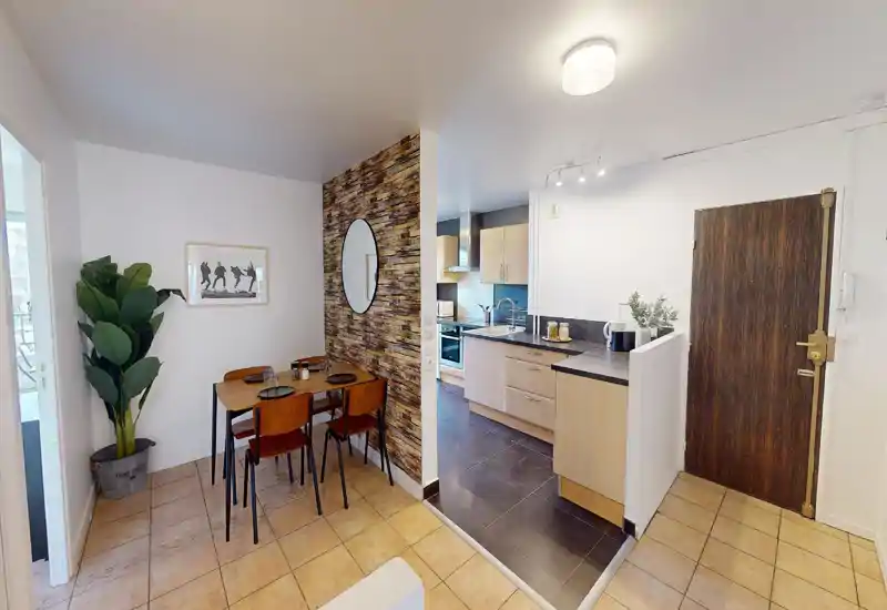 Open-plan living area with a small dining table and a view into the modern kitchen; warm decorative accents include a large potted plant and framed art.