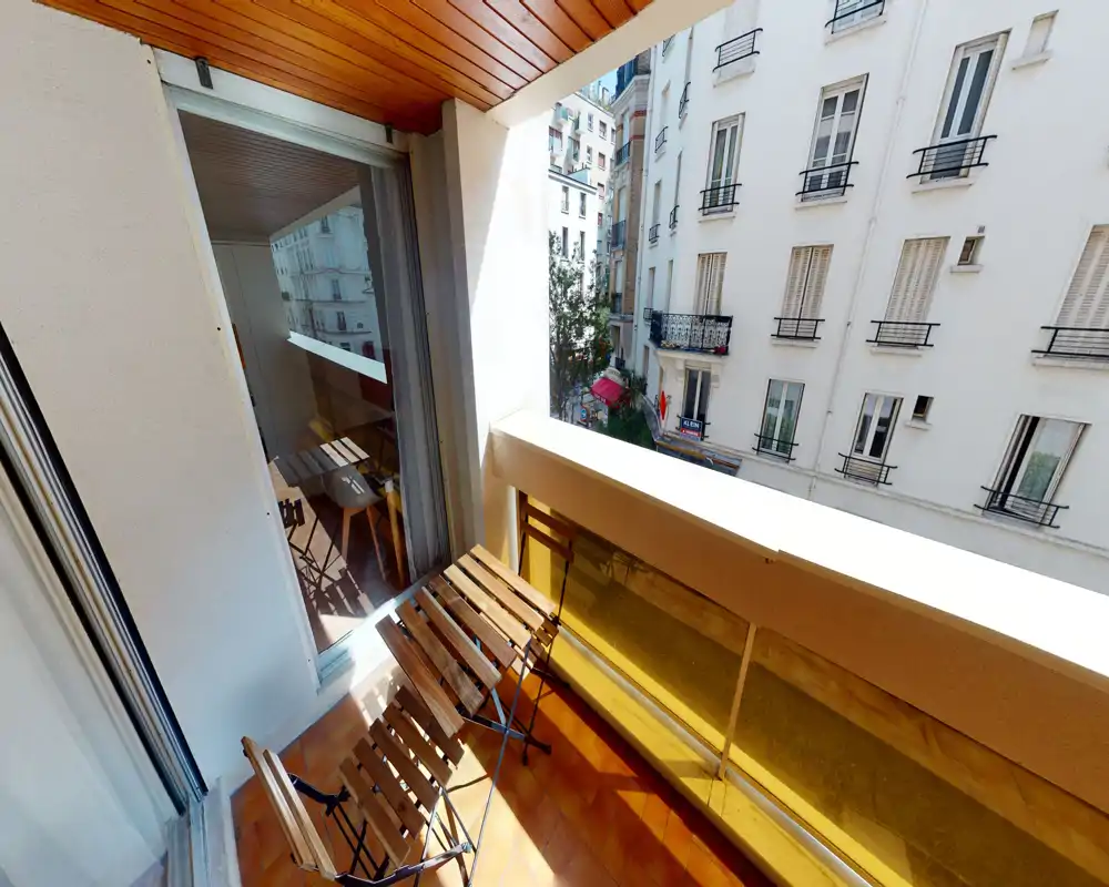 Petit balcon ensoleillé avec une table bistrot en bois et deux chaises pliantes, donnant sur les façades des immeubles voisins — idéal pour le café du matin.