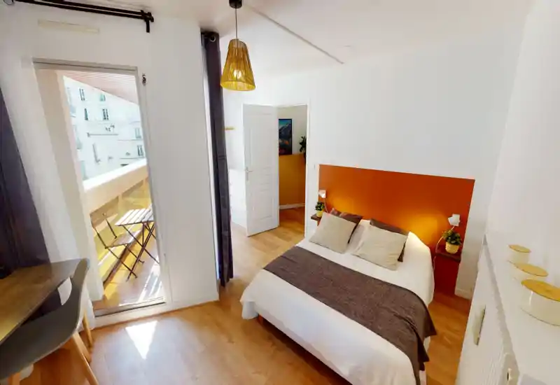 Chambre lumineuse et cosy avec lit double, tête de lit orange chaleureuse et accès au balcon apportant une belle lumière naturelle.