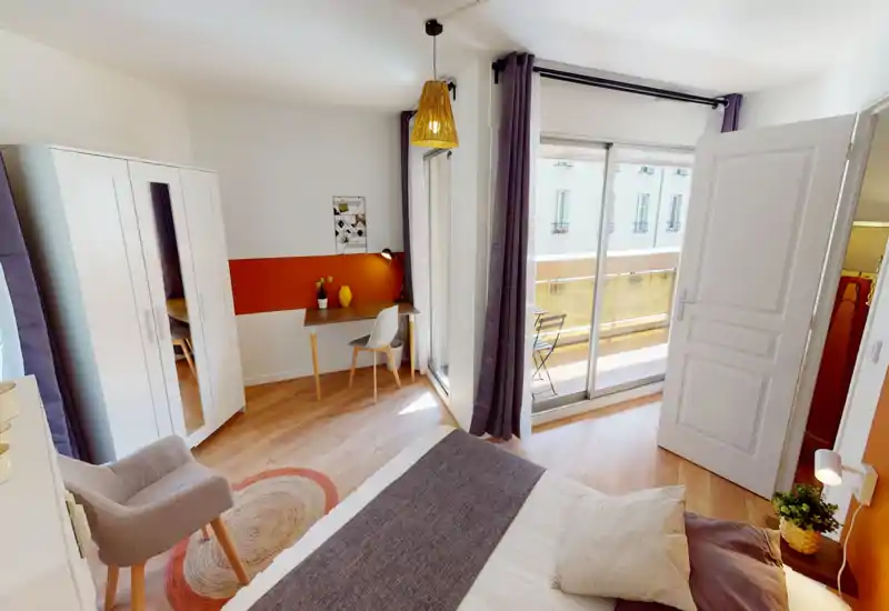 Chambre moderne et lumineuse avec un lit double confortable, parquet, armoire avec miroir et un petit bureau près du balcon. Grandes baies vitrées apportant beaucoup de lumière naturelle et accès à une petite terrasse.