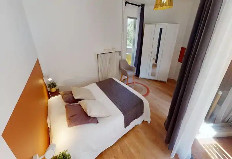 Chambre lumineuse et élégante avec un lit double bien fait, un mur d’accent chaud et la lumière naturelle d’un balcon. Une armoire et un fauteuil apportent confort et harmonie.