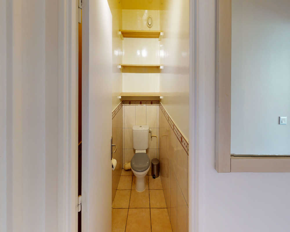 Petit WC fonctionnel avec carrelage au sol et aux murs, étagères au-dessus de la cuvette et petite poubelle. Lumineux, propre et adapté pour de courts séjours.