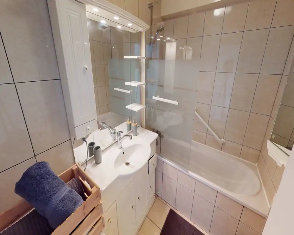 Salle de bains lumineuse et soignée avec lavabo, armoire-miroir et baignoire munie d'une paroi de douche en verre; carrelage neutre et rangements pratiques.