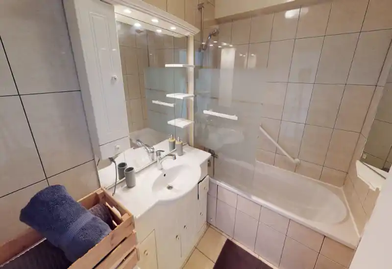 Well-lit, tidy bathroom with a sink, mirror cabinet and a bathtub with a glass shower screen; neutral tiled walls and practical storage make it functional and inviting.