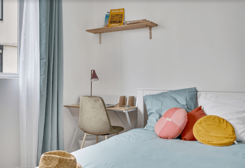 Chambre lumineuse et soignée avec couette bleu clair, coussins ronds décoratifs et bureau près de la fenêtre — idéale pour un séjour cosy.