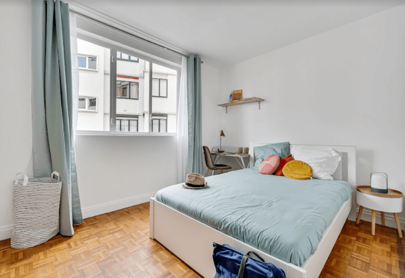 Chambre lumineuse et bien mise en scène avec un lit double, des textiles pastels, une table de chevet et un petit bureau près de la fenêtre. Grande fenêtre apportant une belle lumière naturelle, parquet propre et décoration moderne et minimaliste.