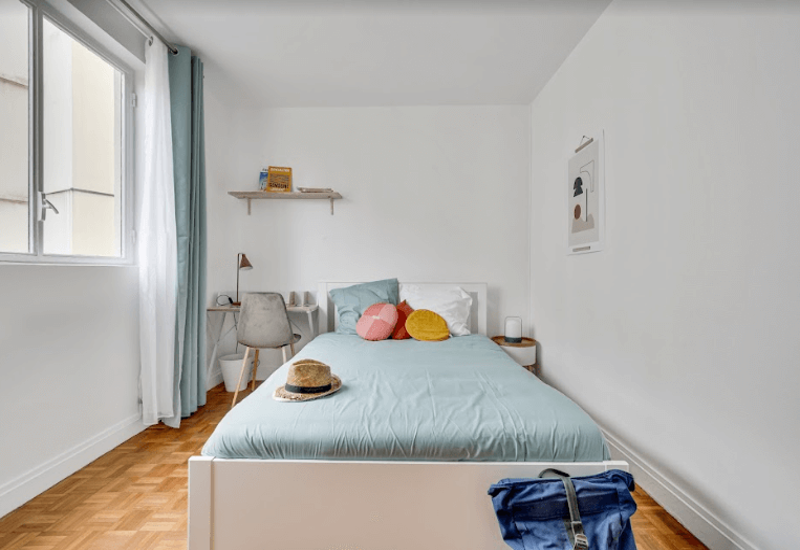 Chambre lumineuse et bien mise en scène avec lit complet, linge de lit pastel et coussins colorés; lumière naturelle par une grande fenêtre et un petit coin bureau à côté du lit.