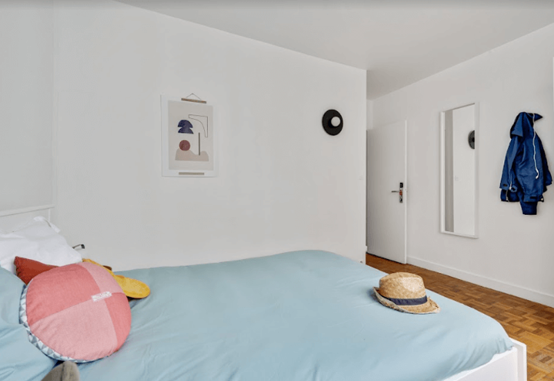Chambre minimaliste et lumineuse avec un grand lit habillé d’une couette bleu pâle, coussins décoratifs et un chapeau de paille posé sur le lit. Murs blancs, parquet et une œuvre murale sobre donnent une atmosphère calme et aérée, idéale pour une annonce.