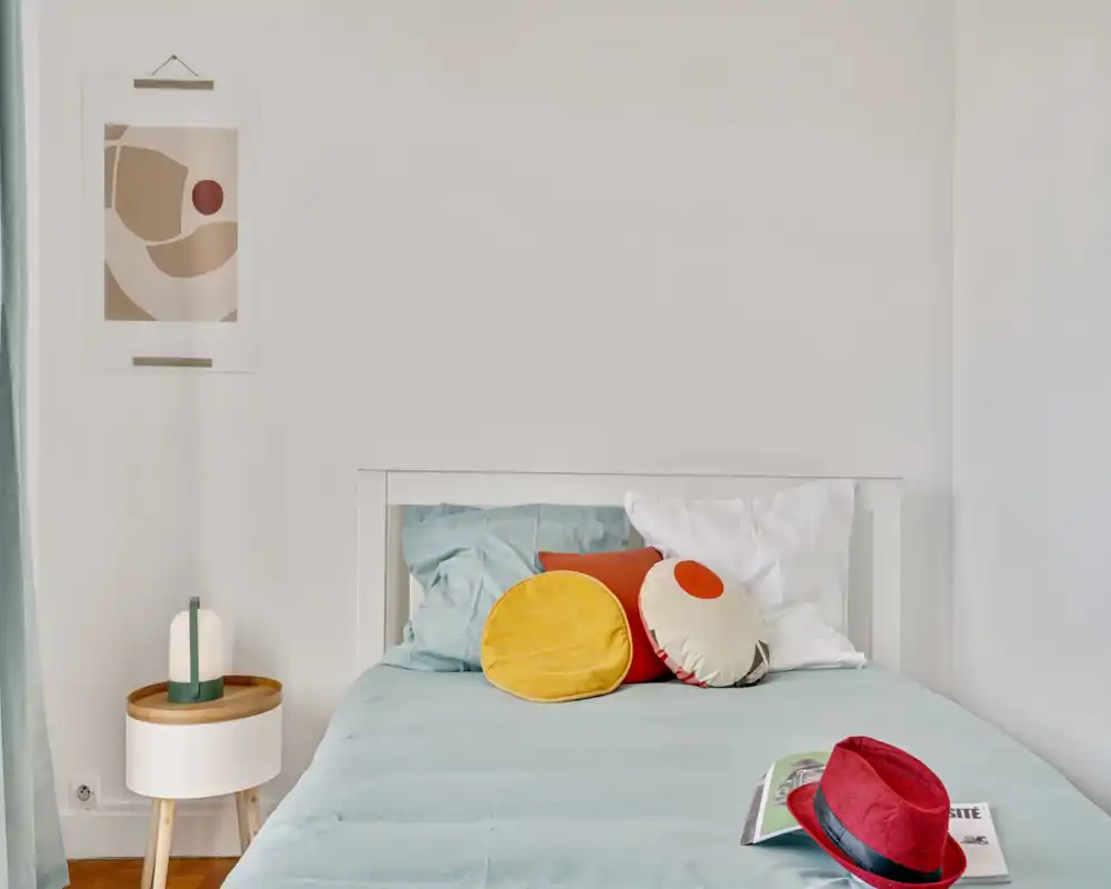 Bright, minimal bedroom with a neatly made bed in soft aqua linens, colorful round cushions and a small bedside table with a lamp—calm, modern and welcoming.