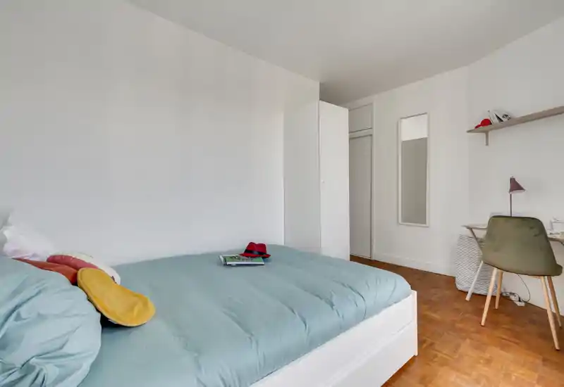 Bright, minimal bedroom with a large bed dressed in light blue bedding, a white wardrobe, wall mirror and a small desk area — airy and simply styled for comfortable stays.