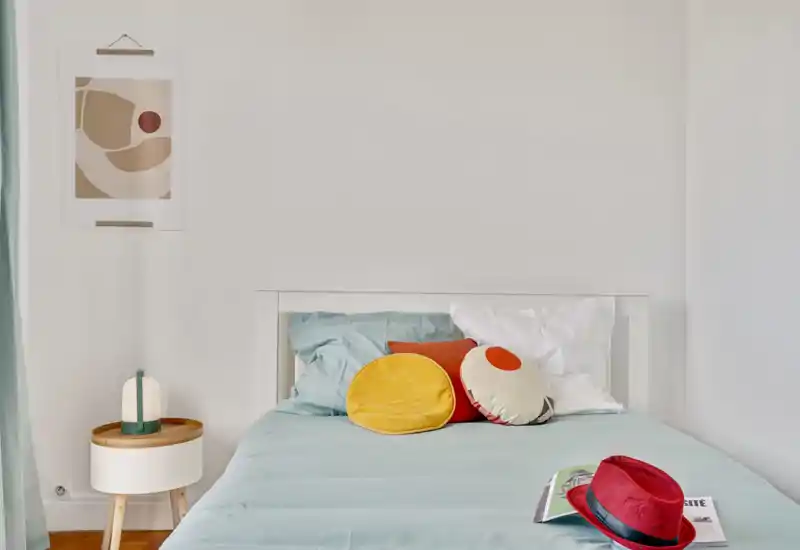 Bright, minimal bedroom with a neatly made bed in soft aqua linens, colorful round cushions and a small bedside table with a lamp—calm, modern and welcoming.