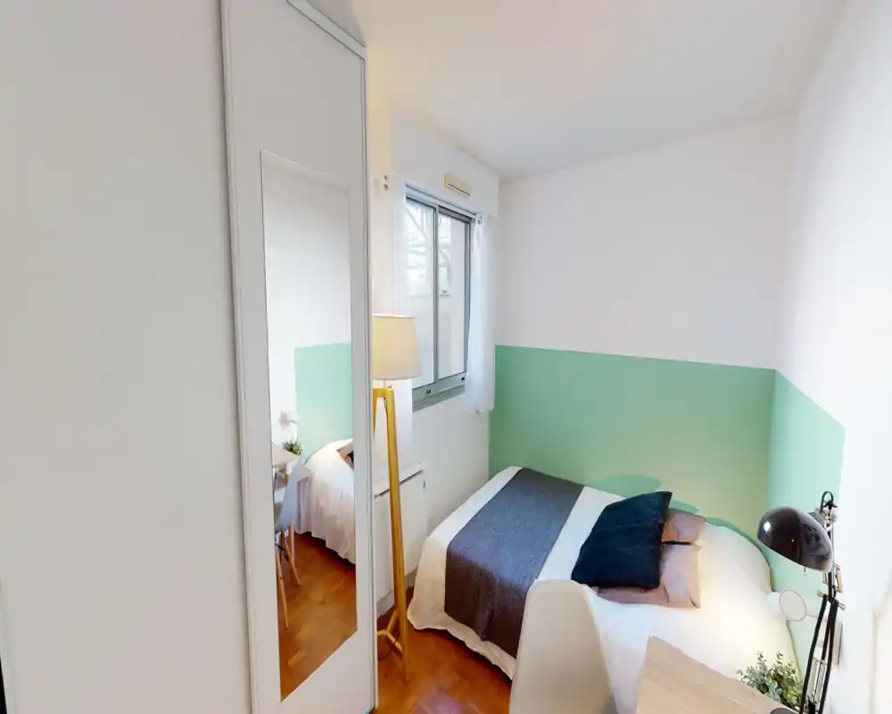 Chambre compacte et lumineuse avec lit en linge blanc, plaid et coussins contrastants, lampadaire et armoire miroir qui renforce la profondeur.