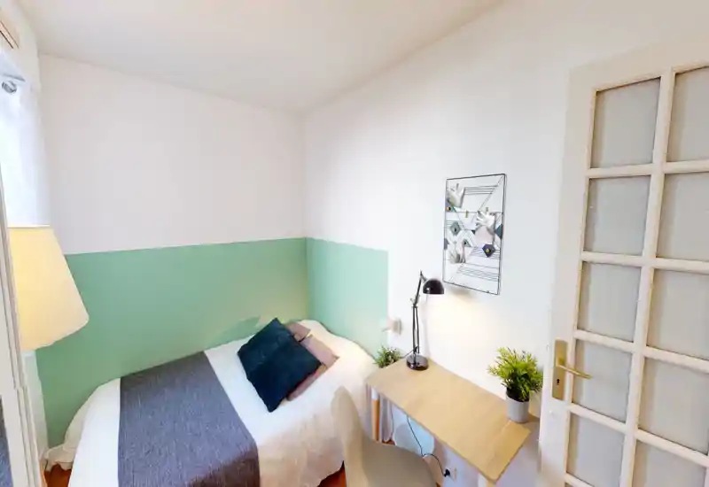 Bright, compact bedroom with a neatly made single/double bed against a two-tone green and white wall, a small wooden desk with a lamp and plants, and soft decorative cushions. The room is well lit and tastefully staged for rental photos.