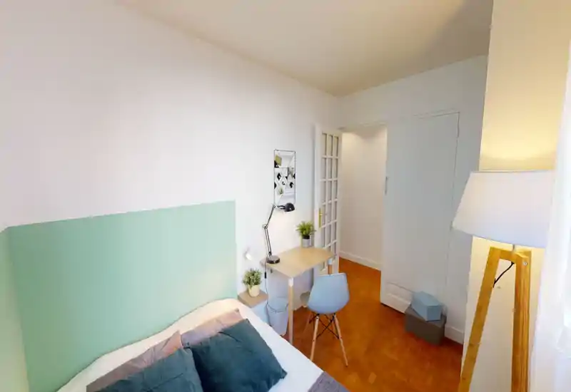Bright, compact bedroom with a mint-green headboard, soft pillows and a small workspace. The room feels cozy and well-lit, ideal for a single traveler or student.