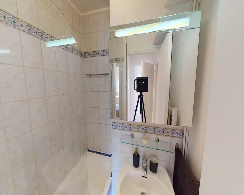 Salle de bains carrelée et compacte avec baignoire, armoire-miroir murale et lavabo avec accessoires. Éclairage neutre et frise décorative bleue pour un espace propre et fonctionnel.