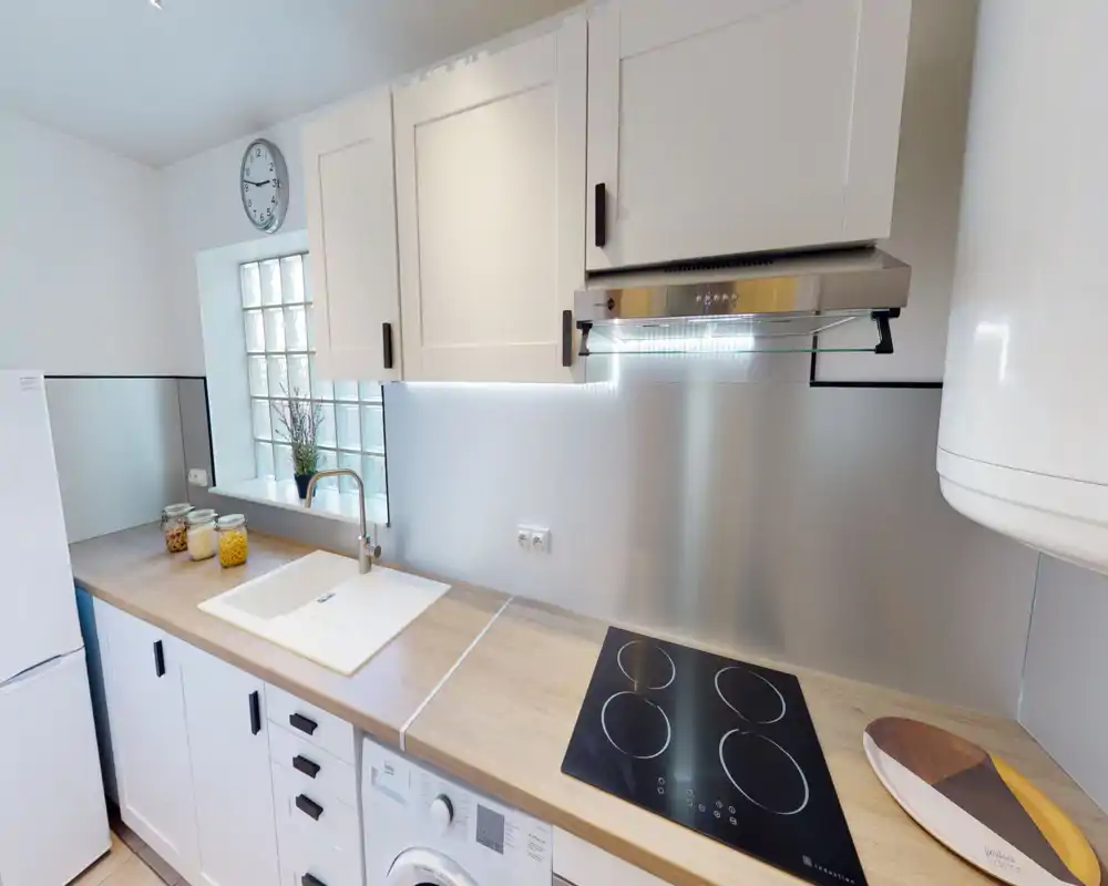 Cuisine compacte et moderne avec placards blancs, plan de travail en bois, plaque à induction intégrée et évier encastré. Style lumineux et soigné avec rangements pratiques et petites touches décoratives.