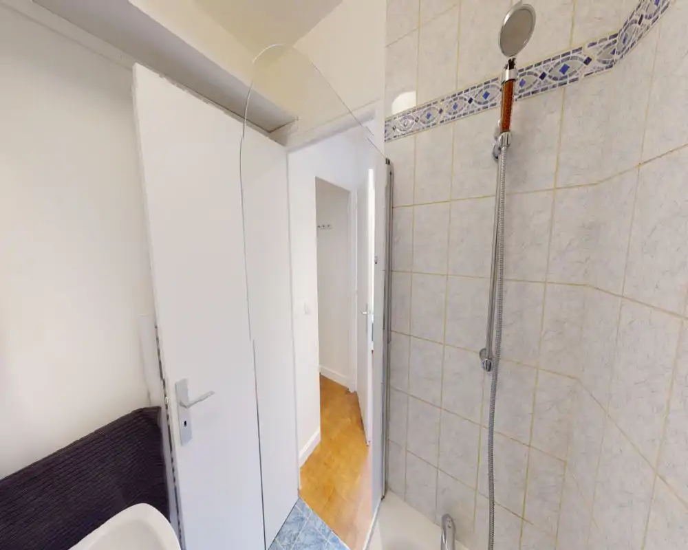 Salle de bains compacte avec coin douche carrelé et pommeau de douche à main. La photo montre la zone de la douche, les murs carrelés et une partie de la porte donnant sur un couloir en parquet.